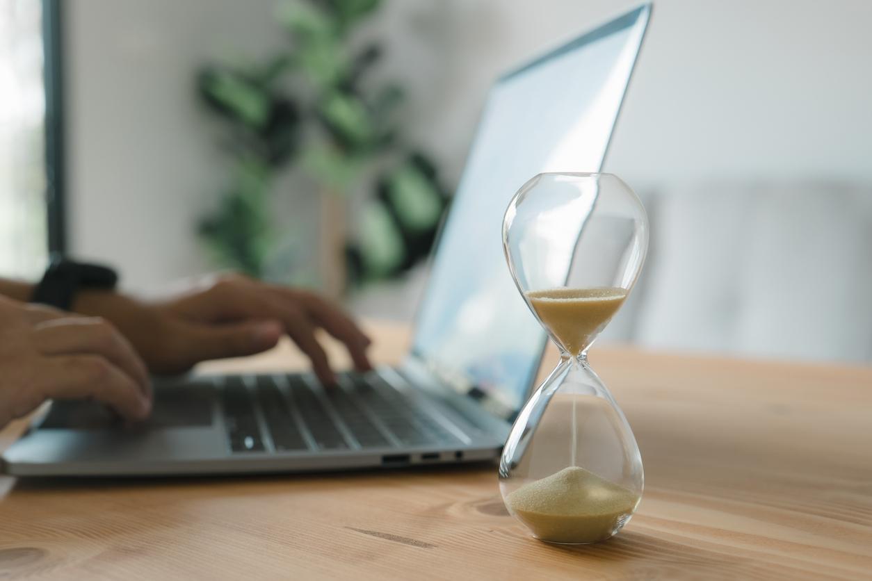 An hourglass with sand running out in front of a person using a laptop